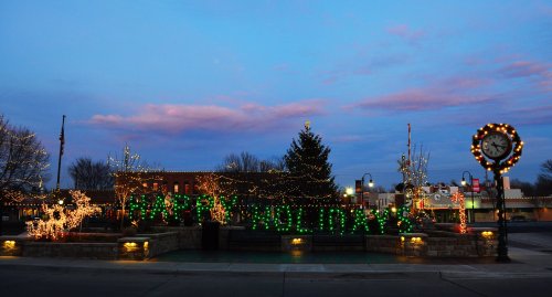 A festive town square decorated with holiday lights, a Christmas tree, and a clock at dusk.
