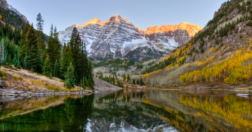 A serene lake reflects snow-capped mountains and autumn trees, illuminated by the warm glow of sunset.