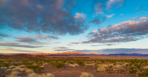 A vast desert landscape under a colorful sky with clouds, featuring distant mountains and sparse vegetation.