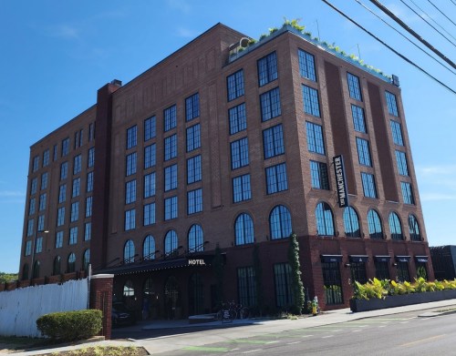 A modern brick hotel building with large windows and greenery on the roof, set against a clear blue sky.