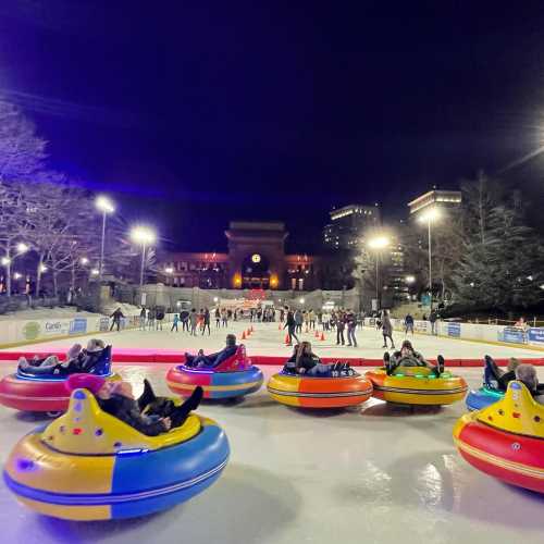 Colorful bumper cars on an ice rink at night, with people enjoying the activity and city lights in the background.