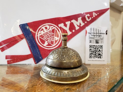 A vintage bell sits on a display with a YMCA pennant and a hotel brochure featuring a QR code.