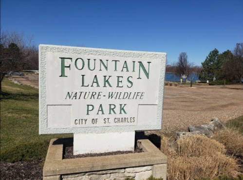 Sign for Fountain Lakes Nature-Wildlife Park in St. Charles, with a clear blue sky and lake in the background.