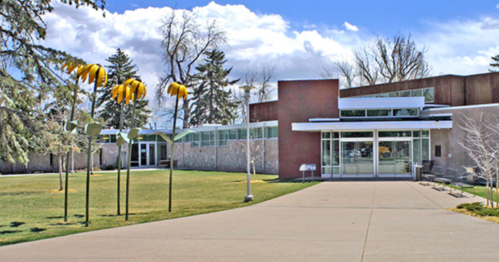 Modern building with large windows, surrounded by green grass and tall, whimsical flower sculptures under a blue sky.
