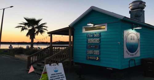 A colorful food stand by the water at sunset, featuring signs for fish tacos, burgers, and snow cones.