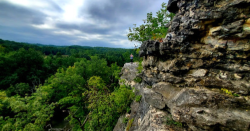 A person stands on a rocky ledge overlooking a lush green landscape under a cloudy sky.