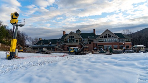 A large building with a green roof surrounded by snow, under a cloudy sky, with snowmaking equipment in the foreground.