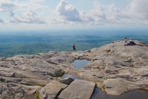 A panoramic view from a rocky mountain summit, with hikers enjoying the scenery and clouds in the blue sky above.