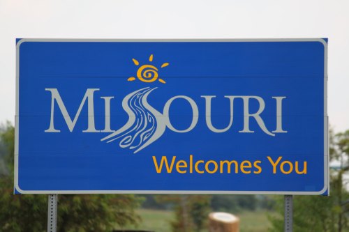 Blue welcome sign for Missouri featuring a sun and flowing river design. Text reads "Missouri Welcomes You."