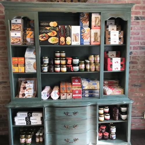 A vintage green cabinet filled with various packaged foods, jars, and snacks on display against a brick wall.