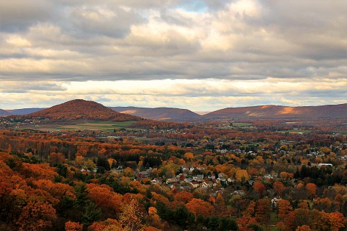 A scenic view of a valley with autumn foliage, rolling hills, and a cloudy sky in the background.