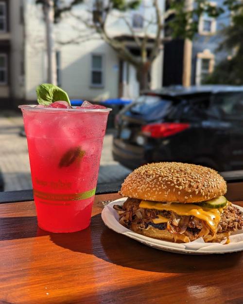 A sesame seed burger with cheese and onions next to a pink drink with mint, set against a street view.