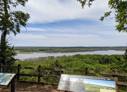 A scenic view of a river surrounded by lush greenery under a partly cloudy sky, with informational signs in the foreground.