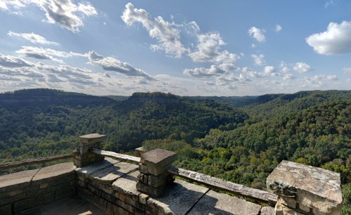 A scenic view of lush green hills and valleys under a partly cloudy sky, seen from a stone overlook.