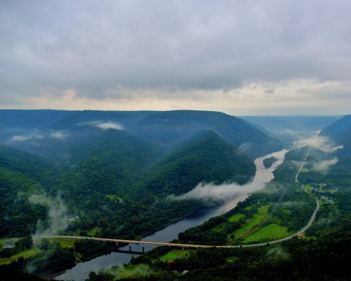 Aerial view of a river winding through lush green mountains, with mist hovering over the landscape under a cloudy sky.