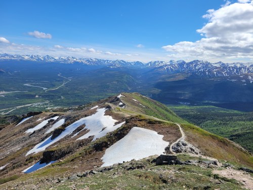 A panoramic view of snow-capped mountains and a lush green valley under a bright blue sky with scattered clouds.