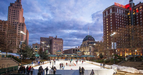 A bustling outdoor ice skating rink surrounded by buildings at dusk, with people skating and enjoying the scene.