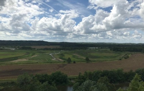 A scenic landscape featuring rolling green fields, a winding river, and a sky filled with fluffy clouds.