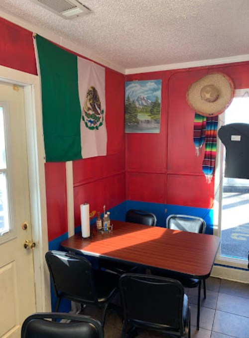 A cozy restaurant corner with a Mexican flag, a painting, sombrero, and colorful decor. Black chairs around a wooden table.