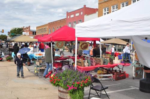 A bustling outdoor market with colorful tents, vendors, and shoppers on a sunny day. Flowers in barrels add charm.