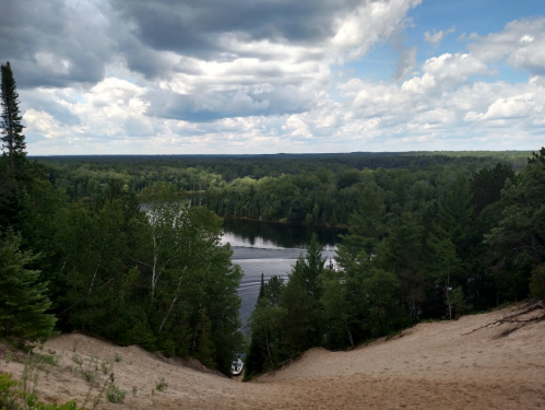 A scenic view of a river surrounded by lush green trees under a cloudy sky, with sandy hills in the foreground.