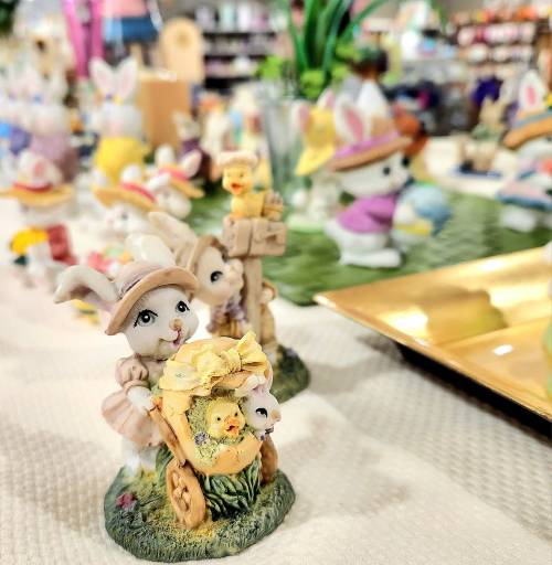 A cute figurine of a bunny in a hat pushing a cart with flowers and chicks, surrounded by colorful Easter decorations.