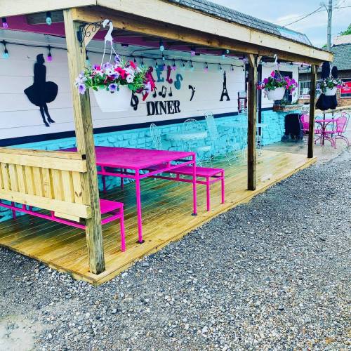 A colorful diner exterior with pink tables, hanging flowers, and a blue wall featuring silhouettes.