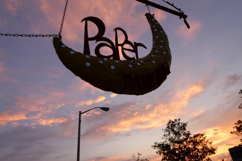A whimsical sign reading "Paper" hangs from a chain against a colorful sunset sky.