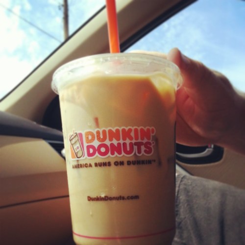 A hand holding a large iced coffee from Dunkin' Donuts inside a car, with a clear blue sky in the background.
