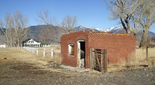A small brick structure stands in a barren landscape with mountains in the background and a distant house.