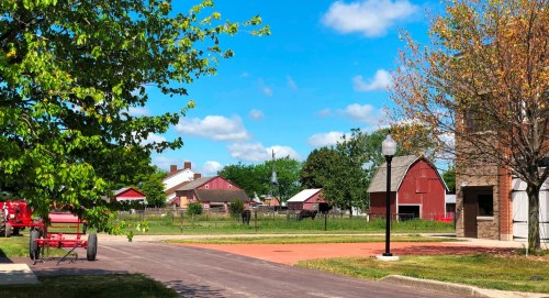 A sunny rural scene featuring red barns, green trees, and a clear blue sky along a paved path.