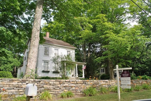 A white two-story house surrounded by lush green trees and a stone wall, with a for-sale sign in front.