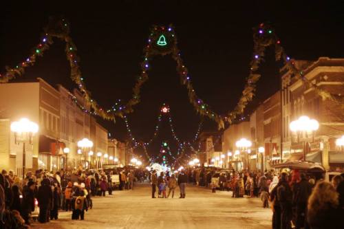A festive street scene at night, lined with holiday lights and decorations, with a crowd gathered along the snowy road.