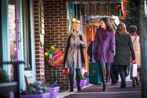 Two women walk along a festive street, smiling and chatting, with holiday decorations and shoppers in the background.