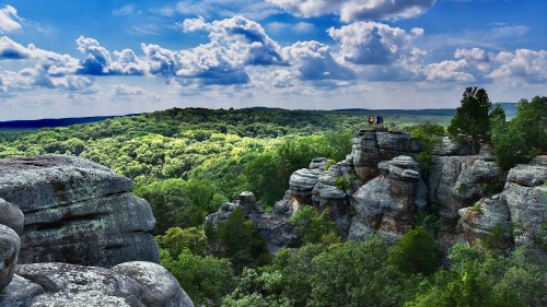 A scenic view of rocky outcrops overlooking a lush green forest under a blue sky with fluffy clouds.