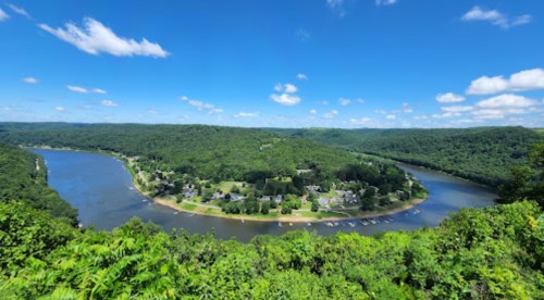 A panoramic view of a river winding through lush green hills under a bright blue sky with scattered clouds.