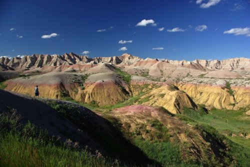 A person stands on a colorful hillside in a vast landscape of layered rock formations under a blue sky.