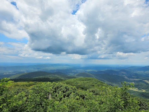 A panoramic view of lush green mountains under a partly cloudy sky, with distant hills and valleys visible.