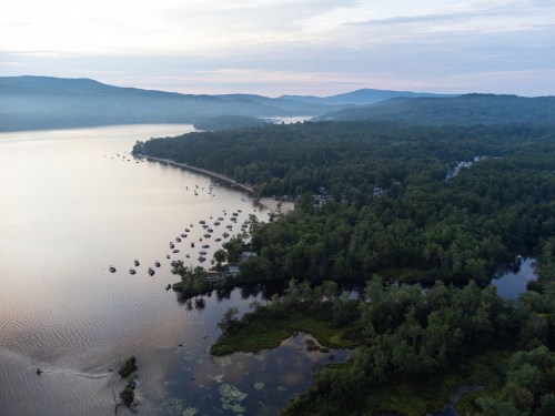 Aerial view of a serene lake surrounded by lush forests and hills, with boats moored along the shore.