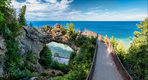 A scenic view of a rocky arch overlooking a vibrant blue lake, with a pathway surrounded by lush greenery.
