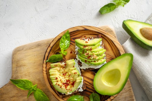 A wooden plate with two avocado toast slices, topped with green apple slices and basil leaves, beside a halved avocado.