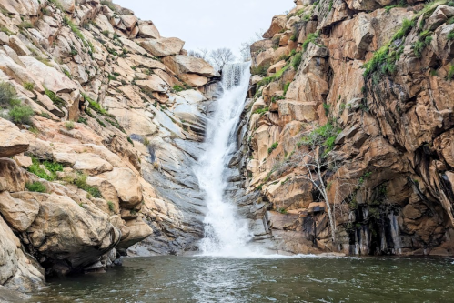 A waterfall cascading down rocky cliffs into a serene pool, surrounded by rugged terrain and sparse vegetation.