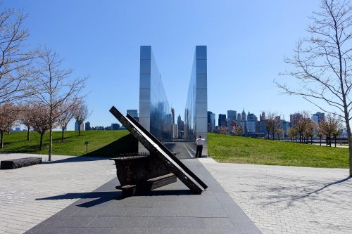 A reflective memorial with a large steel beam in the foreground, set against a clear blue sky and city skyline.