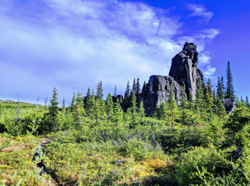 A towering rock formation surrounded by lush green trees under a bright blue sky with scattered clouds.