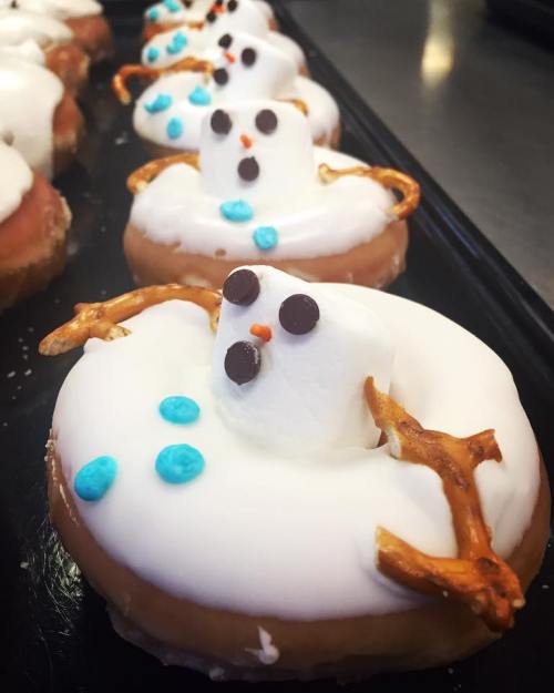 A row of festive donuts decorated as snowmen, featuring marshmallow heads, pretzel arms, and blue icing accents.