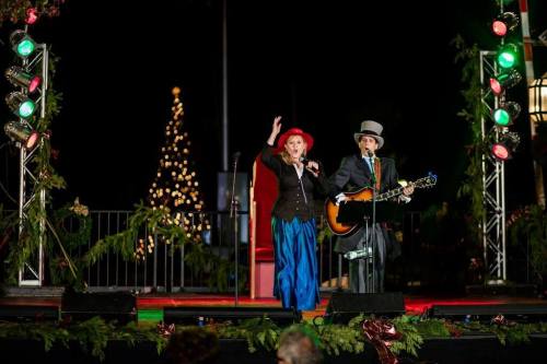 A man and woman perform on stage, dressed in festive attire, with a decorated Christmas tree in the background.