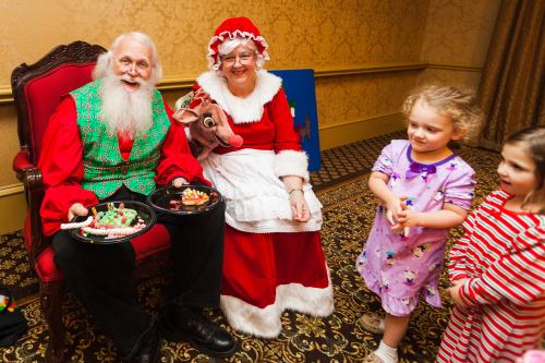 Santa and Mrs. Claus sit together, smiling, with children nearby holding treats in a festive setting.