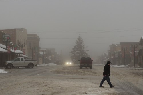 A person walks across a snowy street in a foggy town, with buildings and a Christmas tree in the background.