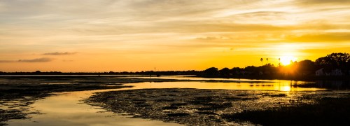A serene sunset over a calm river, with silhouettes of trees and distant buildings against a colorful sky.