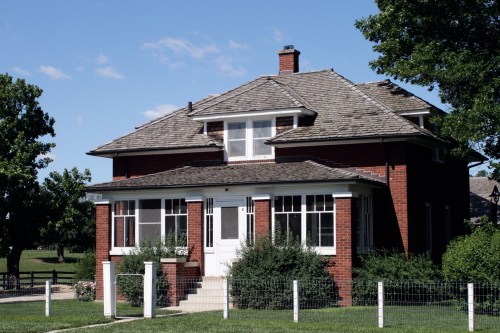 A charming two-story red brick house with a sloped roof, surrounded by greenery and a white picket fence.